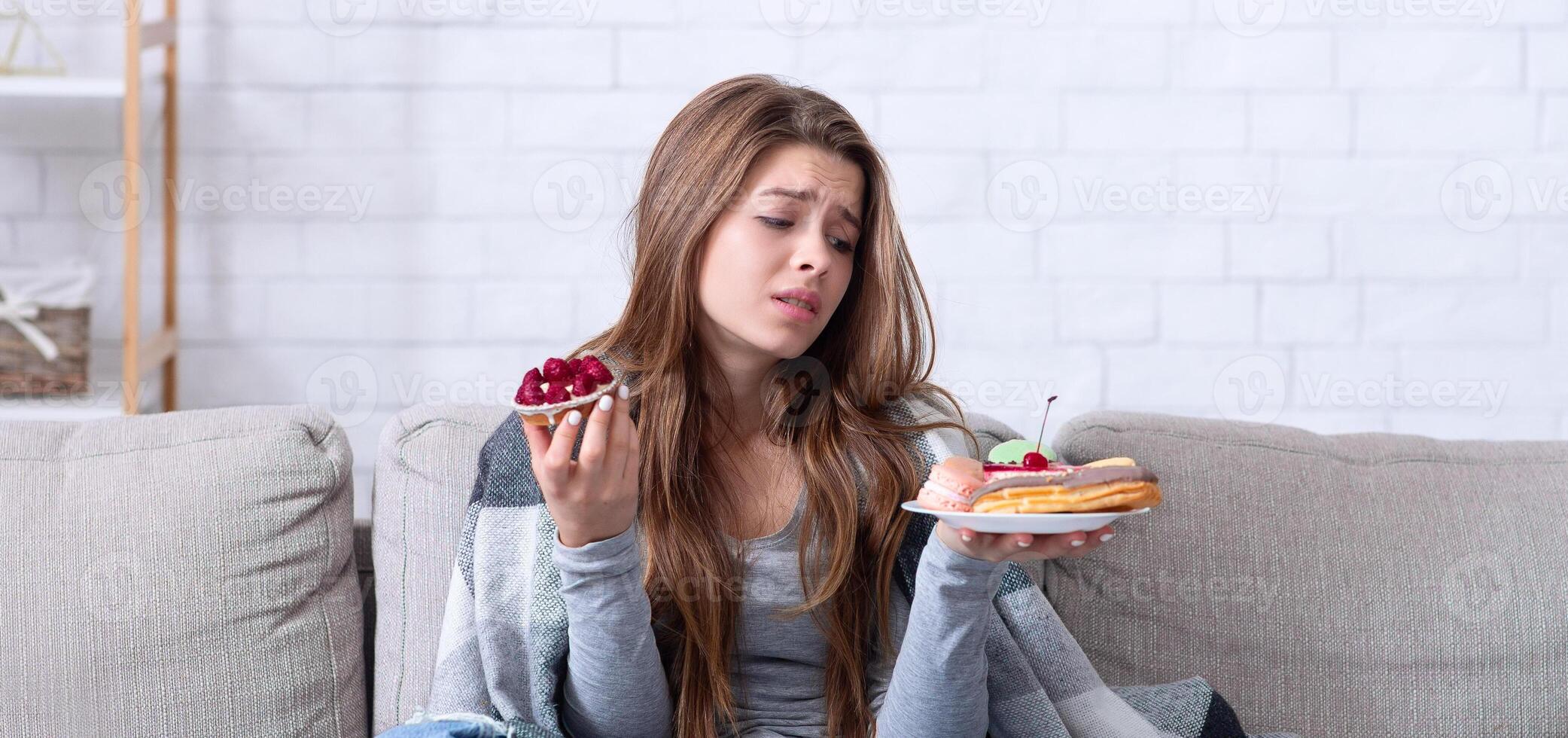 Eating disorders concept. Stressed young woman devouring sweets on couch at home, full length portrait. Unhappy lady coping with negative emotions or problems through unhealthy foods photo