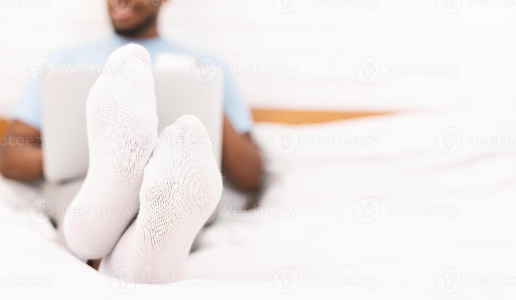 Black guy in white socks working on laptop in bed, feet on foreground, panorama with empty space photo