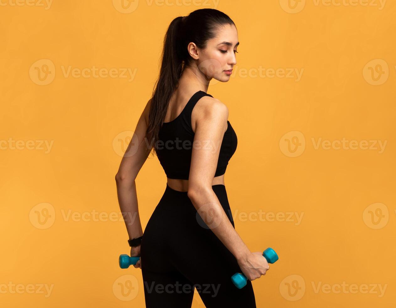 Fitness Model. Portrait Of Confident Lady In Black Sportswear Exercising With Two Dumbbells Posing Isolated Over Orange Background, Turning Back And Looking Down. Studio Shot, Free Space, Banner photo