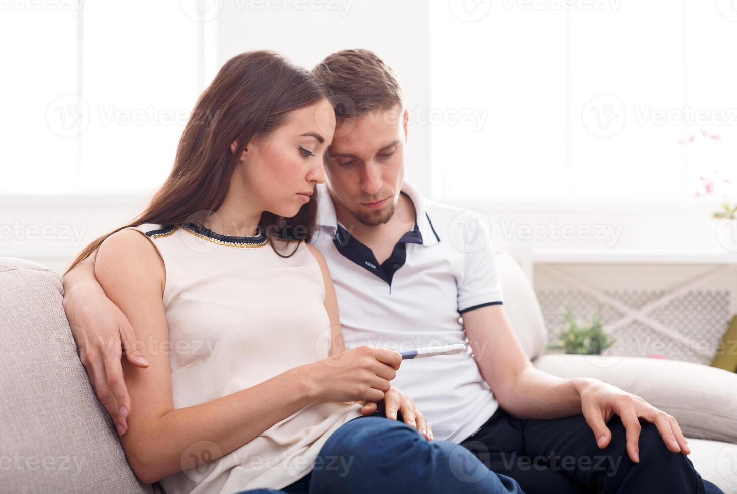 Sad couple after negative pregnancy test result, sitting on couch at home, copy space photo