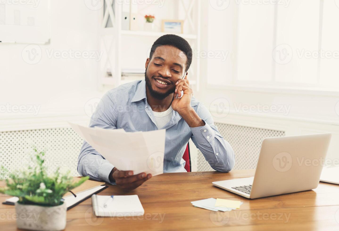 Satisfying call. Young black businessman talking on mobile phone and reading report in modern white office, copy space photo
