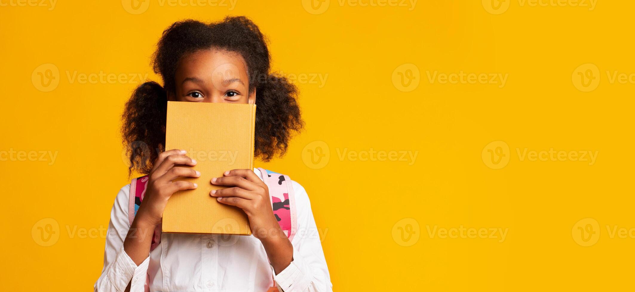 Shy African American Schoolgirl Covering Face With Book On Yellow Background In Studio. Panorama With Empty Space photo