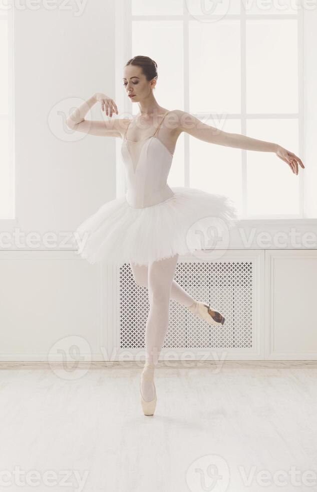 Classical Ballet dancer side view. Beautiful graceful ballerina practice ballet positions in tutu skirt near large window in white light hall. Ballet class training, high-key soft toning. photo
