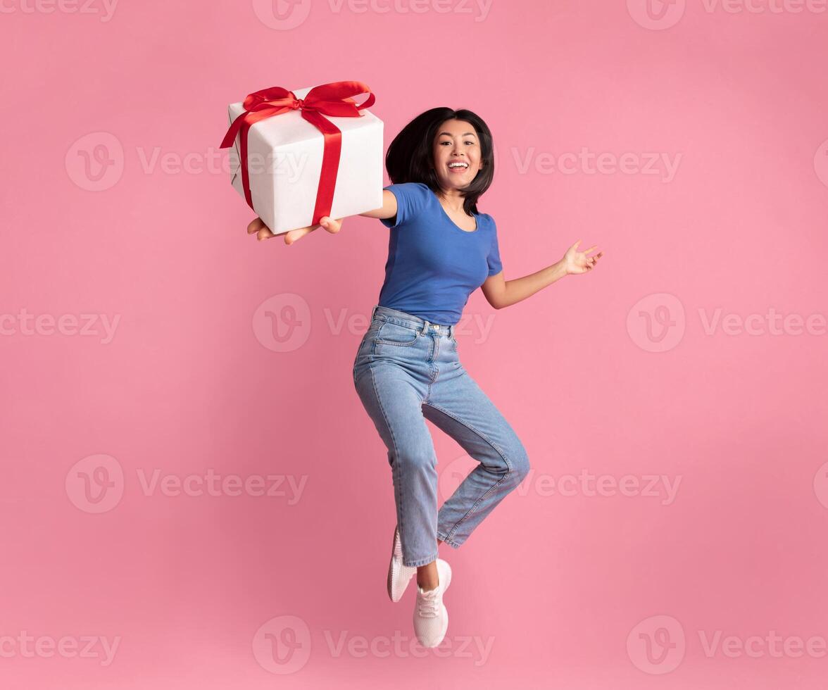 Full length vertical photo of jumping high excited asian lady holding and showing present box close to camera, giving gift isolated over pastel pink studio background. Greeting And Celebration