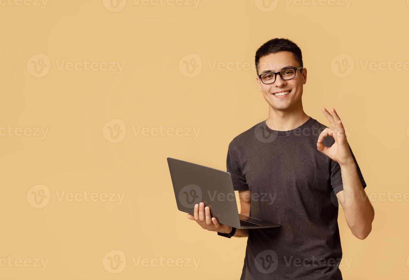 Excellent result, good feedback and win. Smiling young handsome male in glasses expresses emotions of success shows okay gesture and holds laptop, isolated on light background free space, studio shot photo