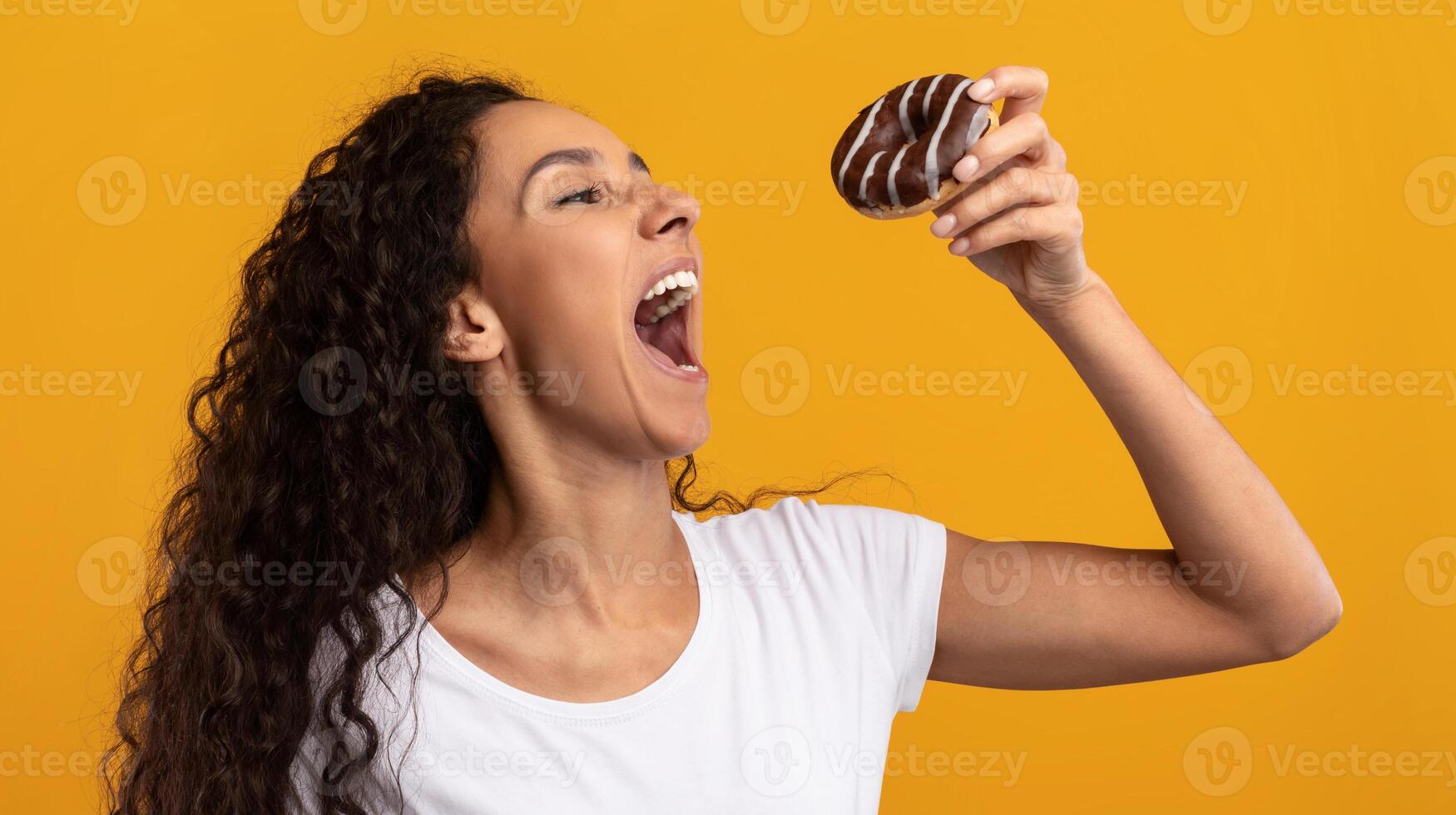 Cheat Meal. Funny Hungry Latin Lady Holding Doughnut Eating Junk Food Posing With Wide Open Mouth Over Yellow Orange Studio Background. Unhealthy Nutrition And Binge Eating Habit Concept photo