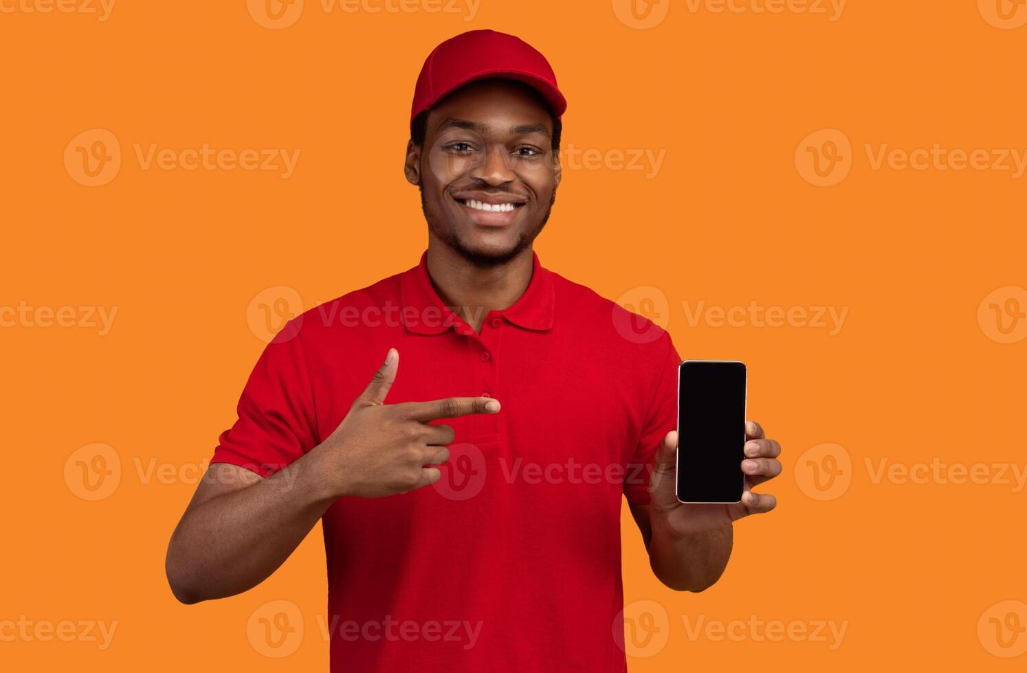 Choose Our Delivery Service. Smiling african american courier holding mobile phone with blank screen for mockup, wearing red cap and uniform, pointing at black empty gadget, orange studio background photo