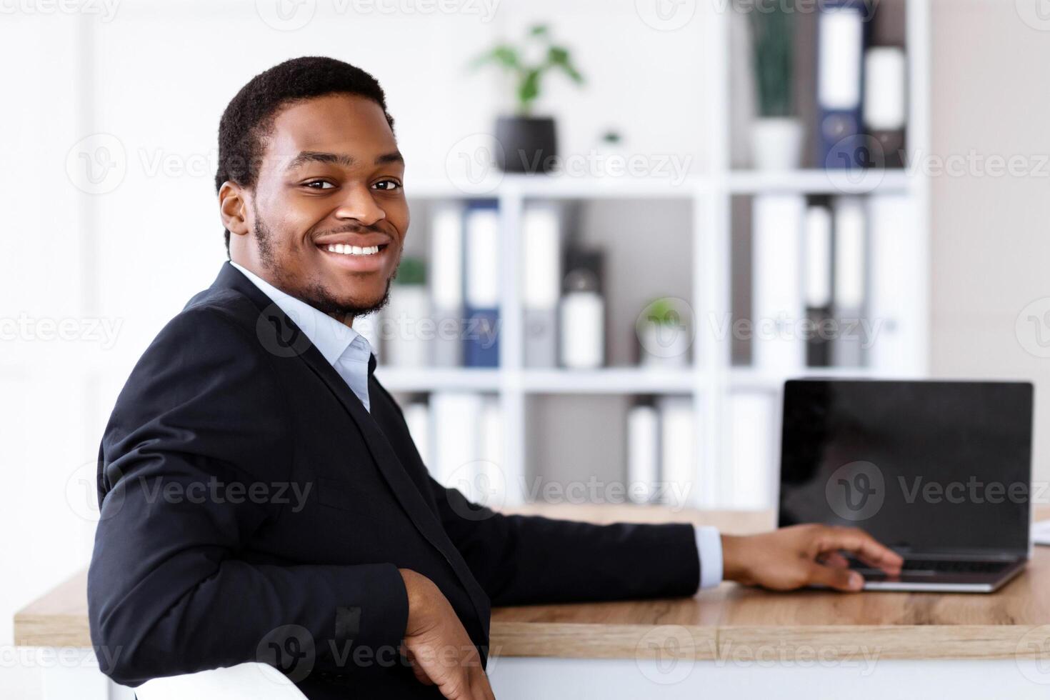 Successful african american businessman posing in his office, using modern laptop with blank screen, smiling at camera, copy space. Cheerful black manager in suit working on computer, mockup photo