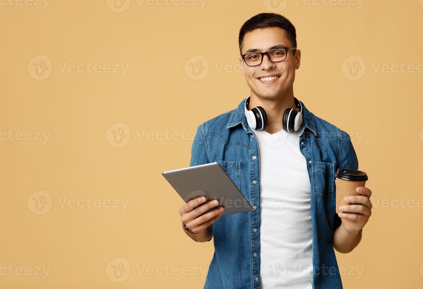 Online creative work and freelance during lockdown coronavirus. Cheerful attractive friendly young guy with glasses and headphones hold digital tablet and coffee takeaway isolated on light background photo