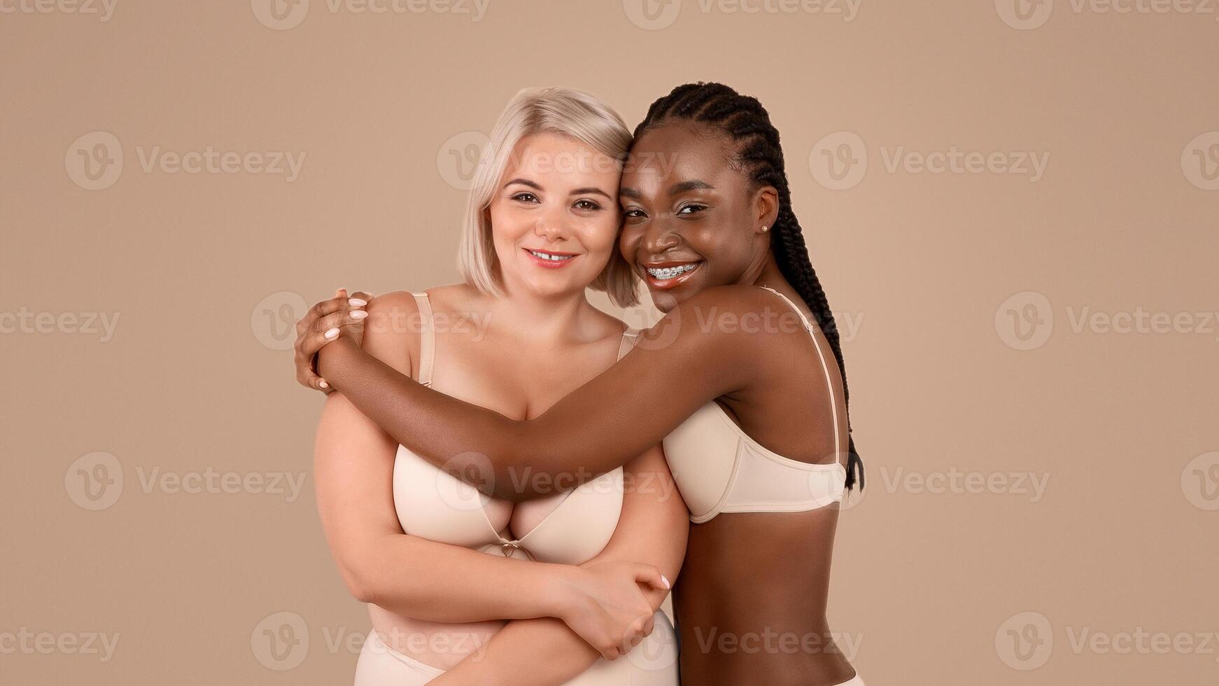 Two body positive ladies in underwear embracing and smiling at camera, black and plus size women supporting each other in self-acceptance, posing in lingerie over beige studio background, copy space photo