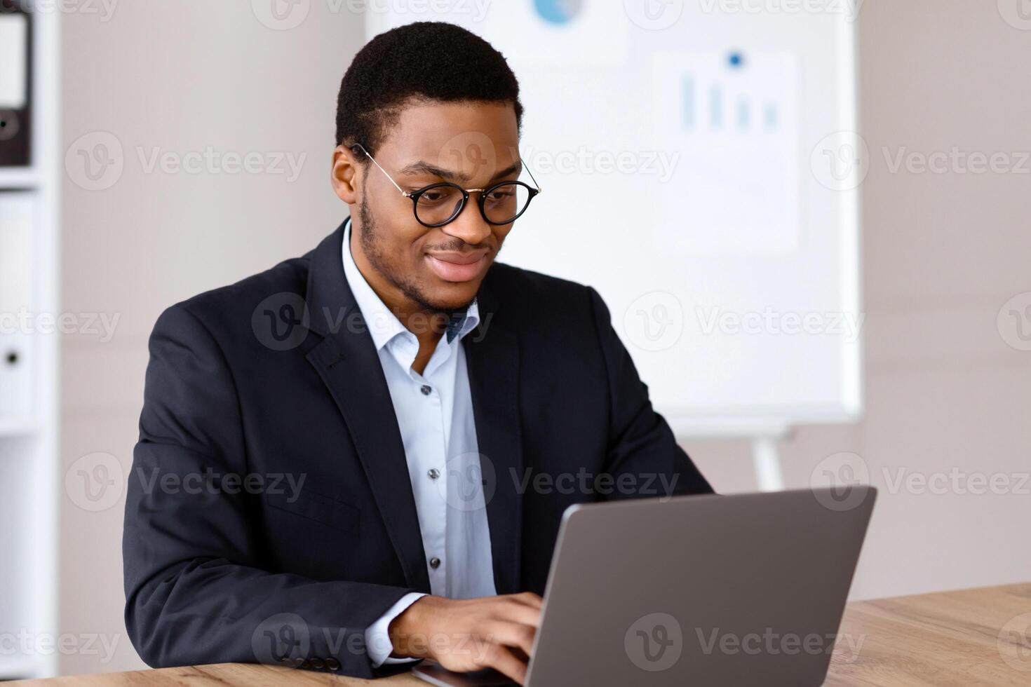 Smiling young black entrepreneur in formal suit working with laptop, wearing glasses, office interior, copy space. Successful millennial african american businessman working on business project photo