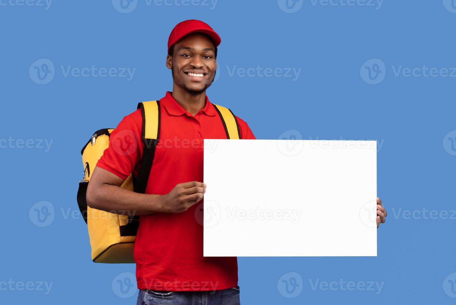 Portrait of cheerful bearded african american deliveryman wearing yellow thermo bag and red uniform holding and showing square empty blank board sign standing isolated on blue studio background photo
