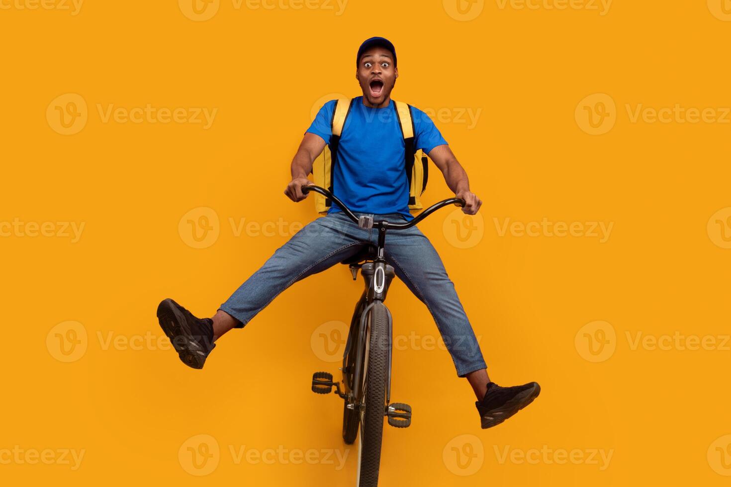 Full length of funny black delivery man in blue cap and uniform riding bicycle, wearing yellow thermo backpack bag isolated over bright orange blue studio background. Fast and quick shipment service photo