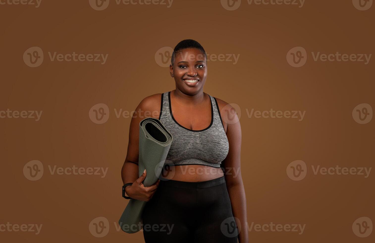 Ready For Training. Portrait of African American chubby woman in bra top and leggings going to gym, holding carrying roll mat standing isolated over brown studio background, looking posing at camera photo