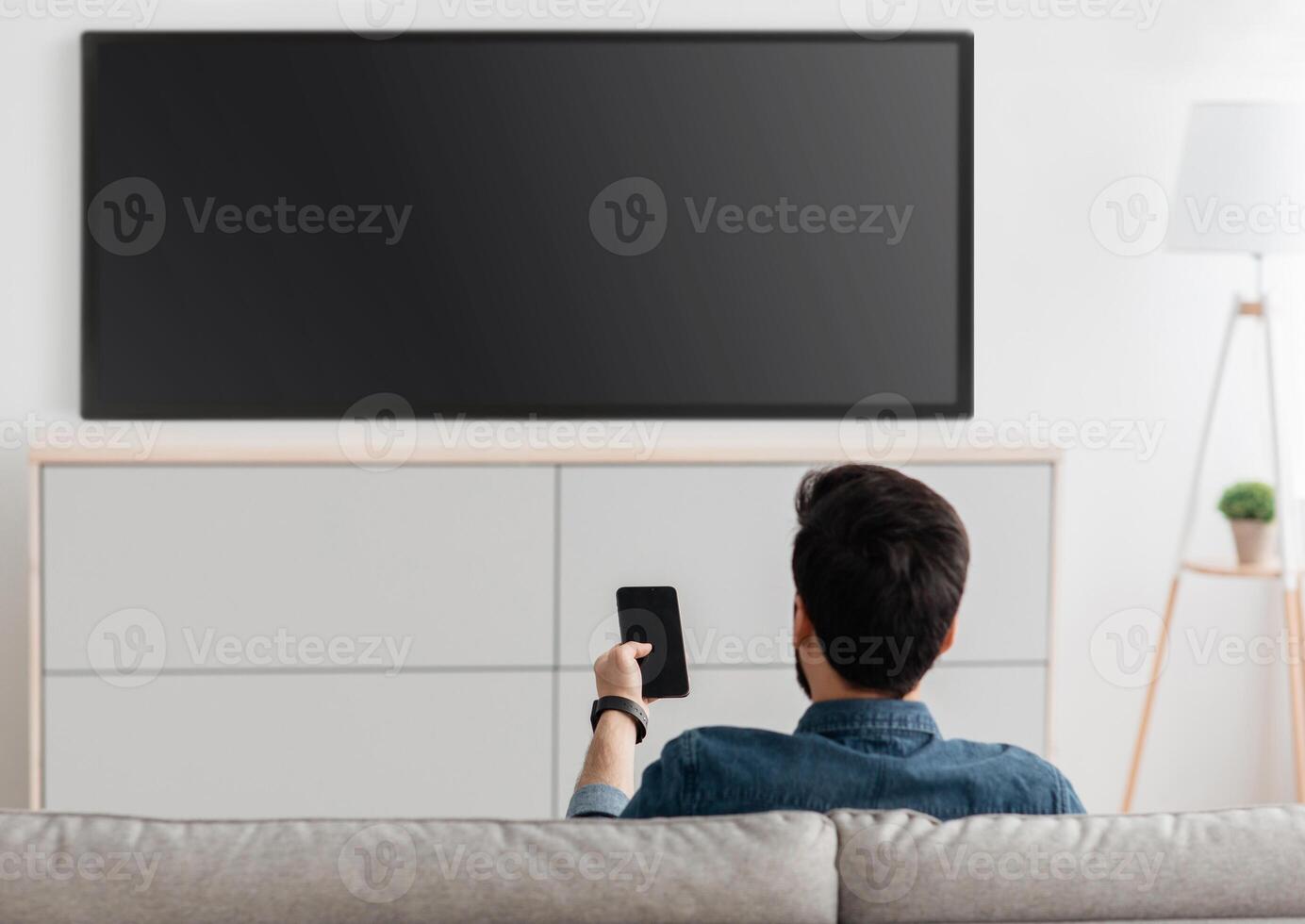 Modern gadgets. Unrecognizable man with smartphone connected to TV, watching at home, back view. Guy transfering from phone to television. Information transfer between devices over wifi. photo