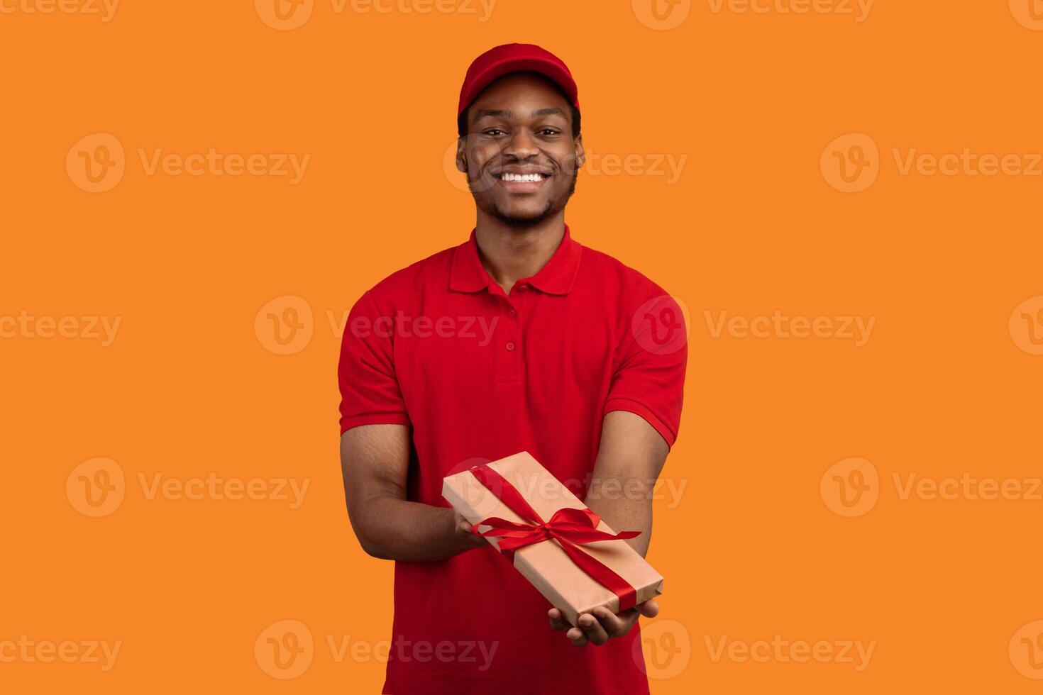 Delivery Service For Holidays. Portrait of smiling cheerful african american courier holding wrapped gift box, giving and shwoing present to camera, isolated on orange studio background photo