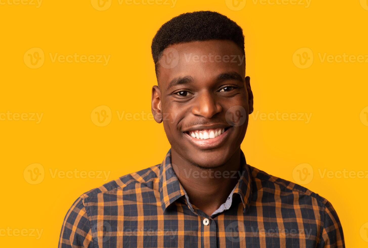 Closeup portrait of positive young African American guy sincerely smiling at camera, cheerful black millennial student guy in checkered shirt standing isolated over yellow background, free space photo