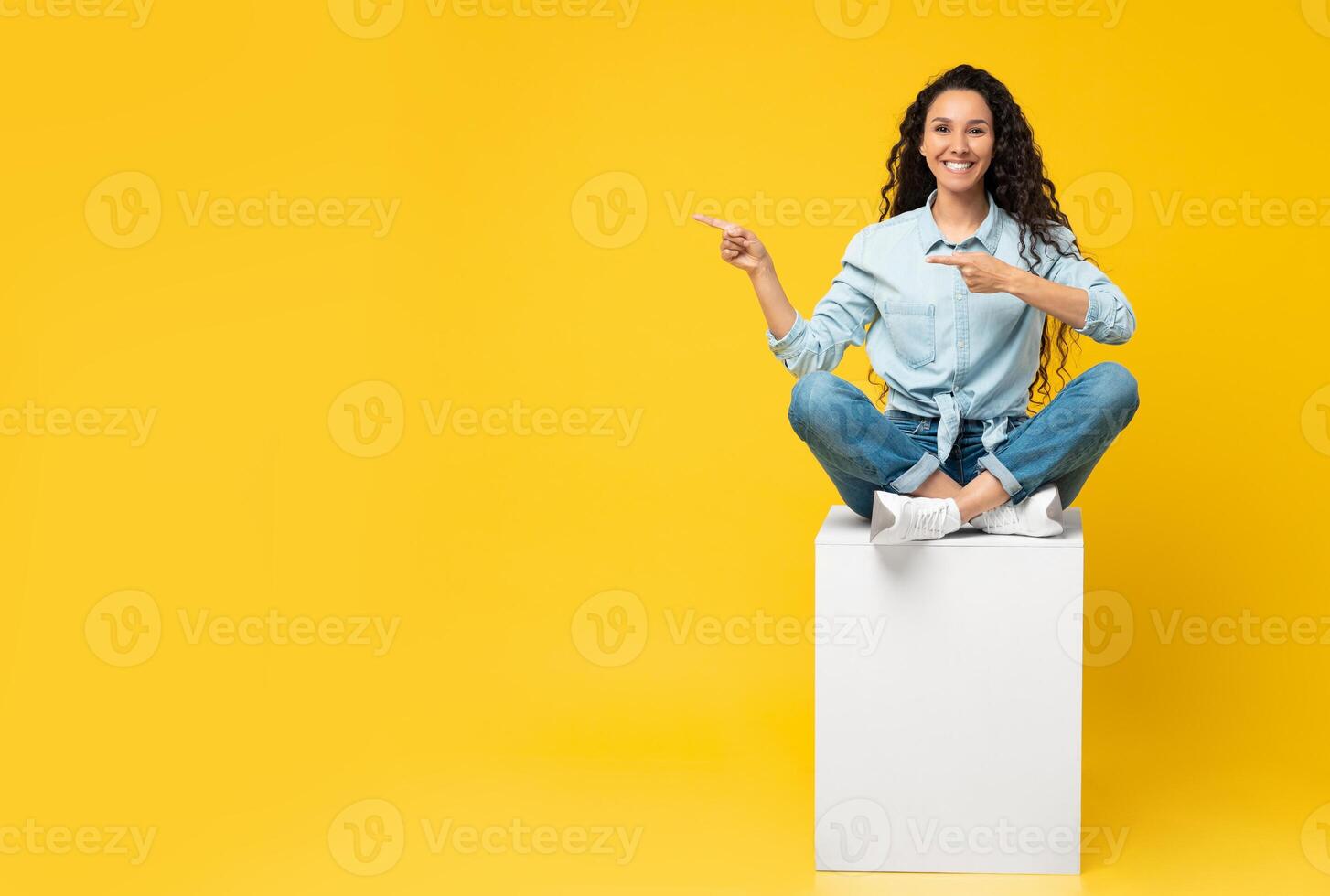 Special Offer. Portrait of excited young lady pointing fingers aside at free copy space sitting on white cube isolated on yellow orange wall. Positive female showing empty mockup place for ad, banner photo