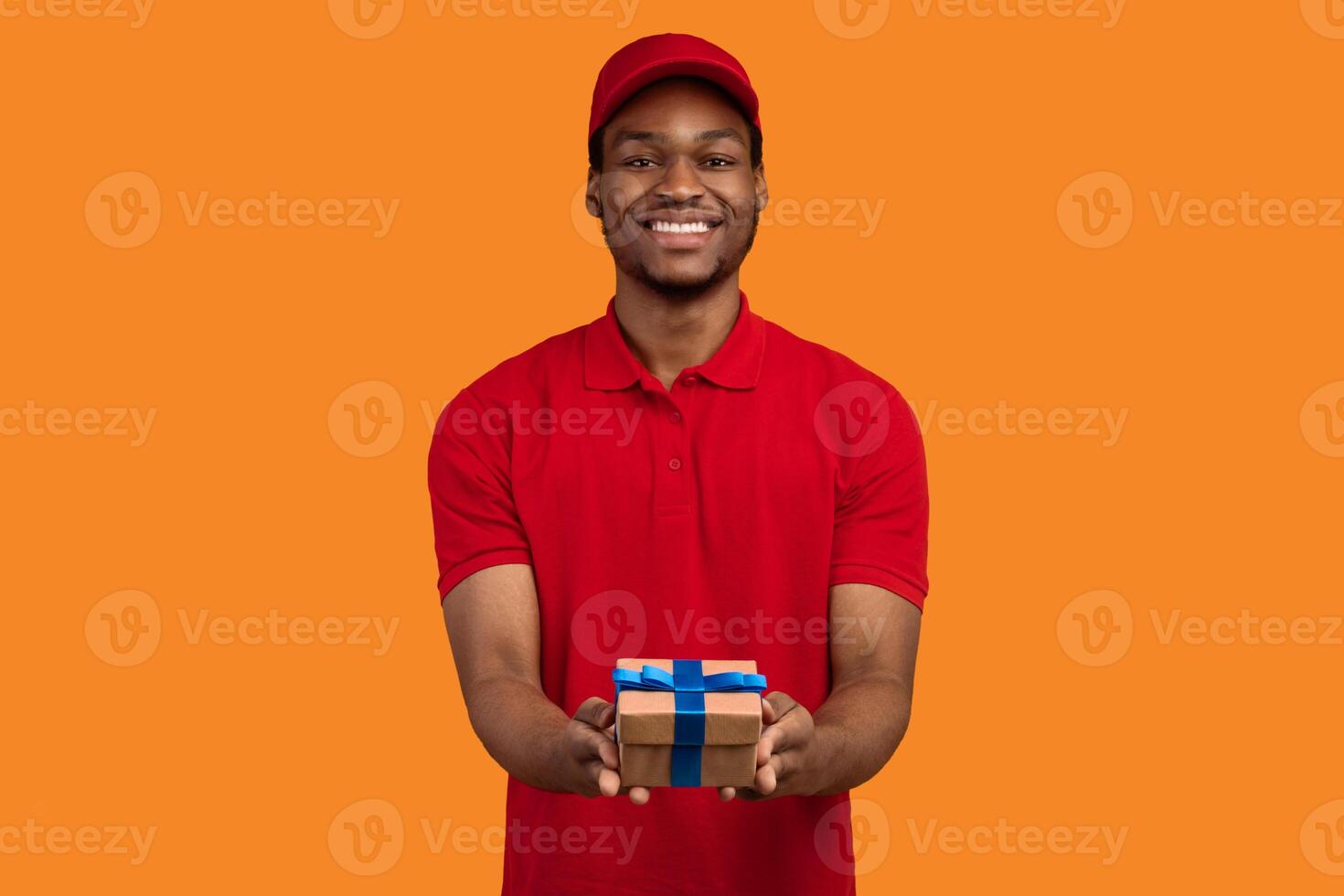 Delivery Service For Holidays. Portrait of smiling cheerful african american courier guy holding wrapped gift box, giving and showing present to camera, isolated over orange studio background photo