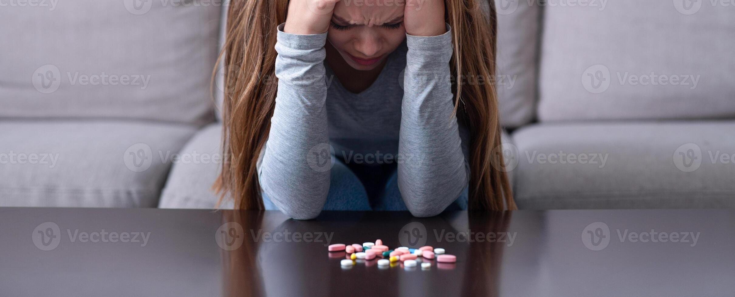 Depressed young woman sitting head in hands near pile of tablets, planning to commit suicide at home. Desperate millennial lady with emotional problems overdosing on meds, killing herself photo