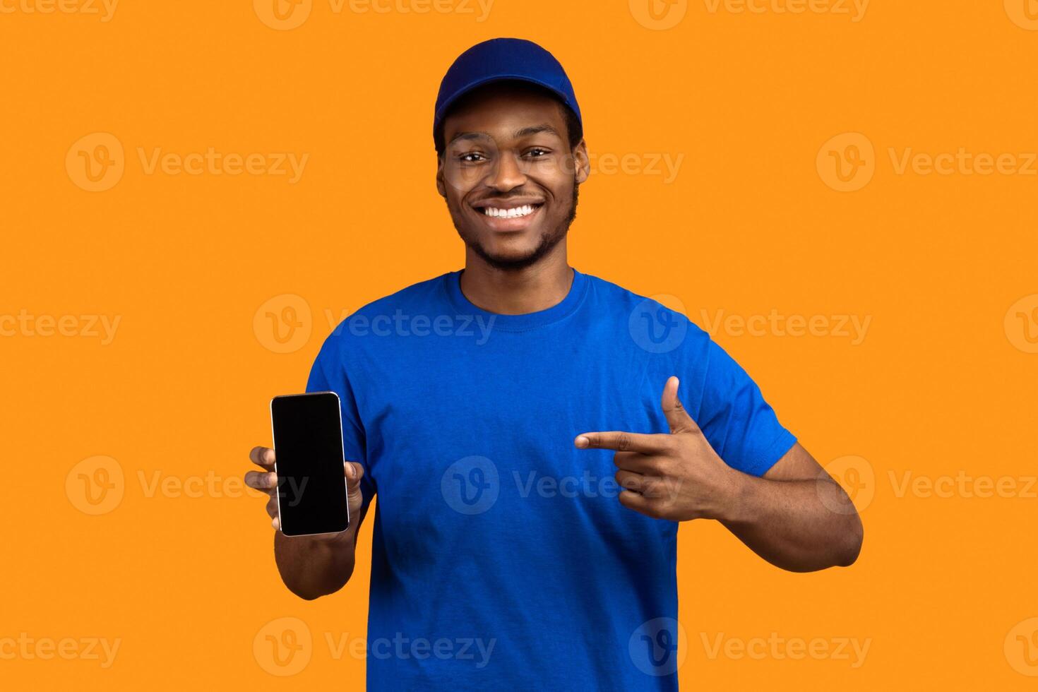 Recommending Service. Smiling african american delivery guy holding smartphone with blank screen for mockup, wearing blue cap and uniform, pointing at black empty monitor isolated on orange background photo