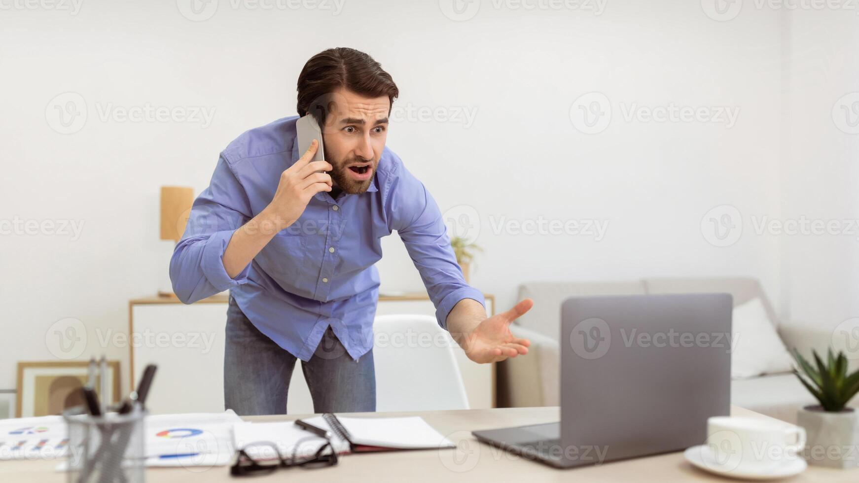Angry Businessman Talking On Phone Looking At Laptop, Having Computer Problem Standing In Office. Communication Issue, Worlplace Problems And Conflicts Concept. Panorama, Selective Focus photo