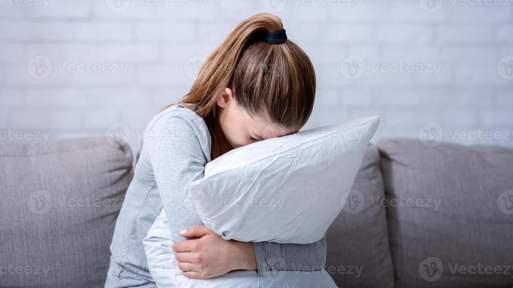 Young woman with depression hugging pillow and crying on sofa at home. Lonely millennial lady feeling stressed and hopeless, suffering from mood disorder or having psychological problem photo