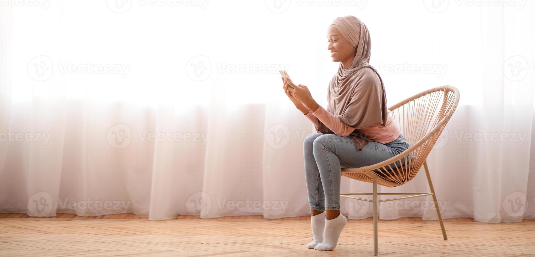 Millennial black lady in hijab using cellphone at home, working or studying remotely, free space. African American woman in Muslim headscarf checking email, sending message, having online call photo