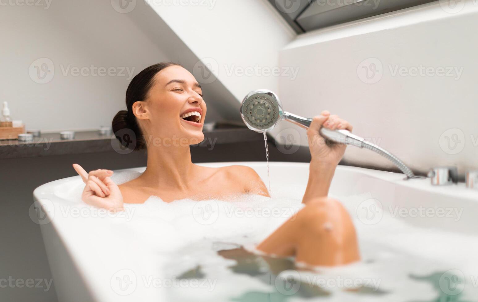 Cheerful Lady Having Fun Taking Bath And Singing Holding Shower Head Like Microphone Sitting In Water With Foam Bubbles In Modern Bathroom At Home. Beauty And Relaxation Leisure photo