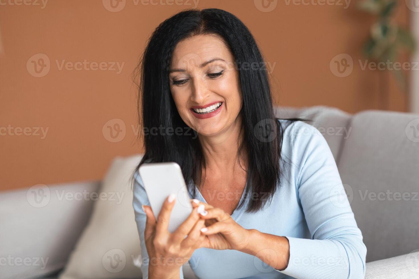 Lifestyle Concept. Portrait of cheerful smiling senior woman holding and using her mobile phone, chatting on internet, texting her children or friends, browsing website, sitting on the sofa at home photo