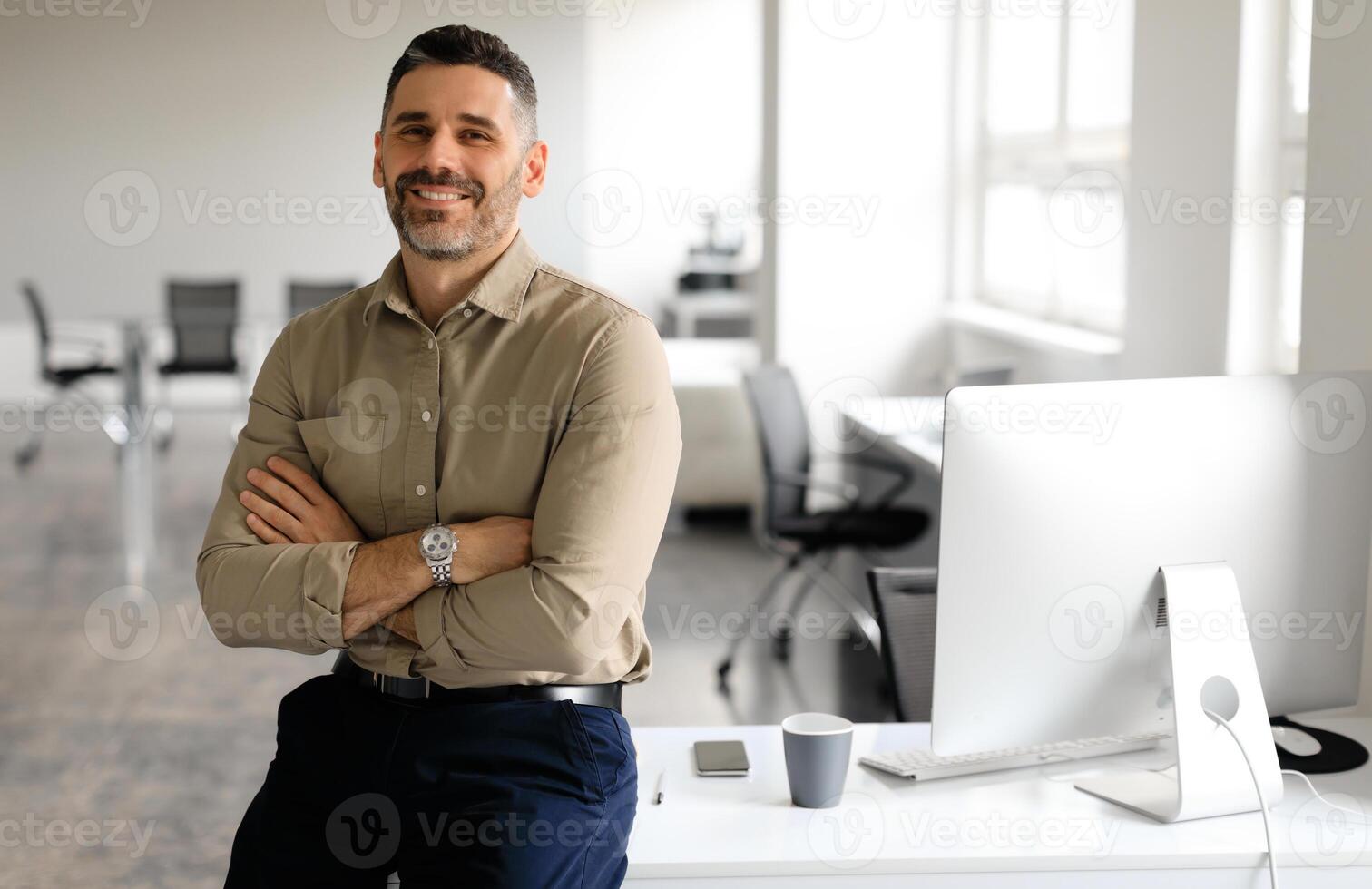 Successful middle aged male entrepreneur sitting with arms crossed at desk in office, looking and smiling at camera, free space. Confident male graphic designer working at company photo