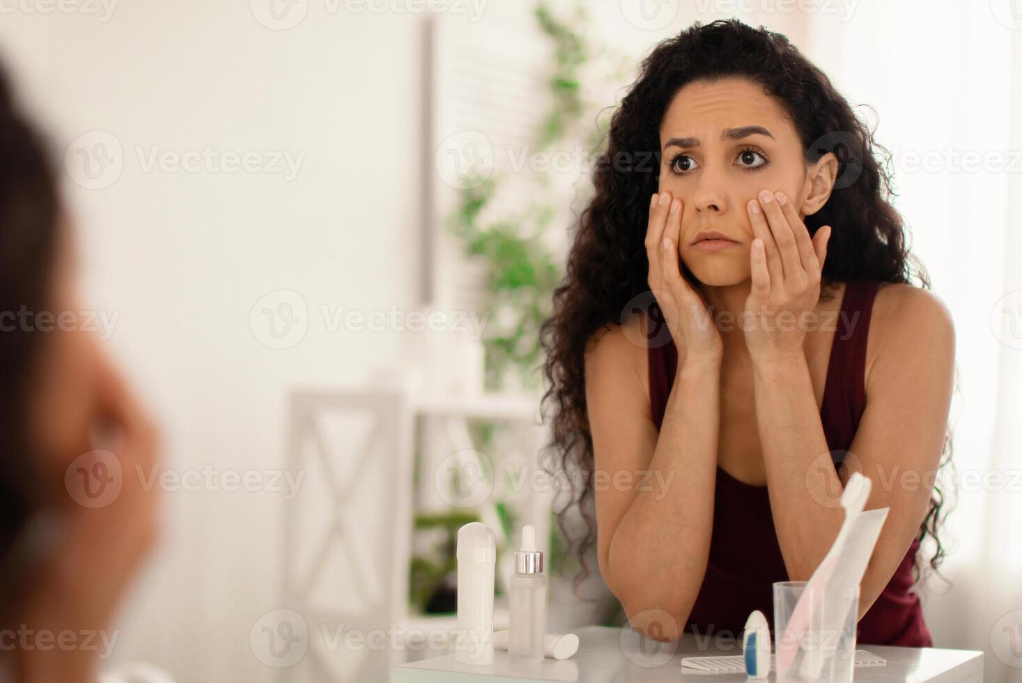 Beauty and skincare. Upset young woman looking in mirror and touching face, examining wrinkles and dark circles under her eyes at home, copy space. Dull tired skin concept photo