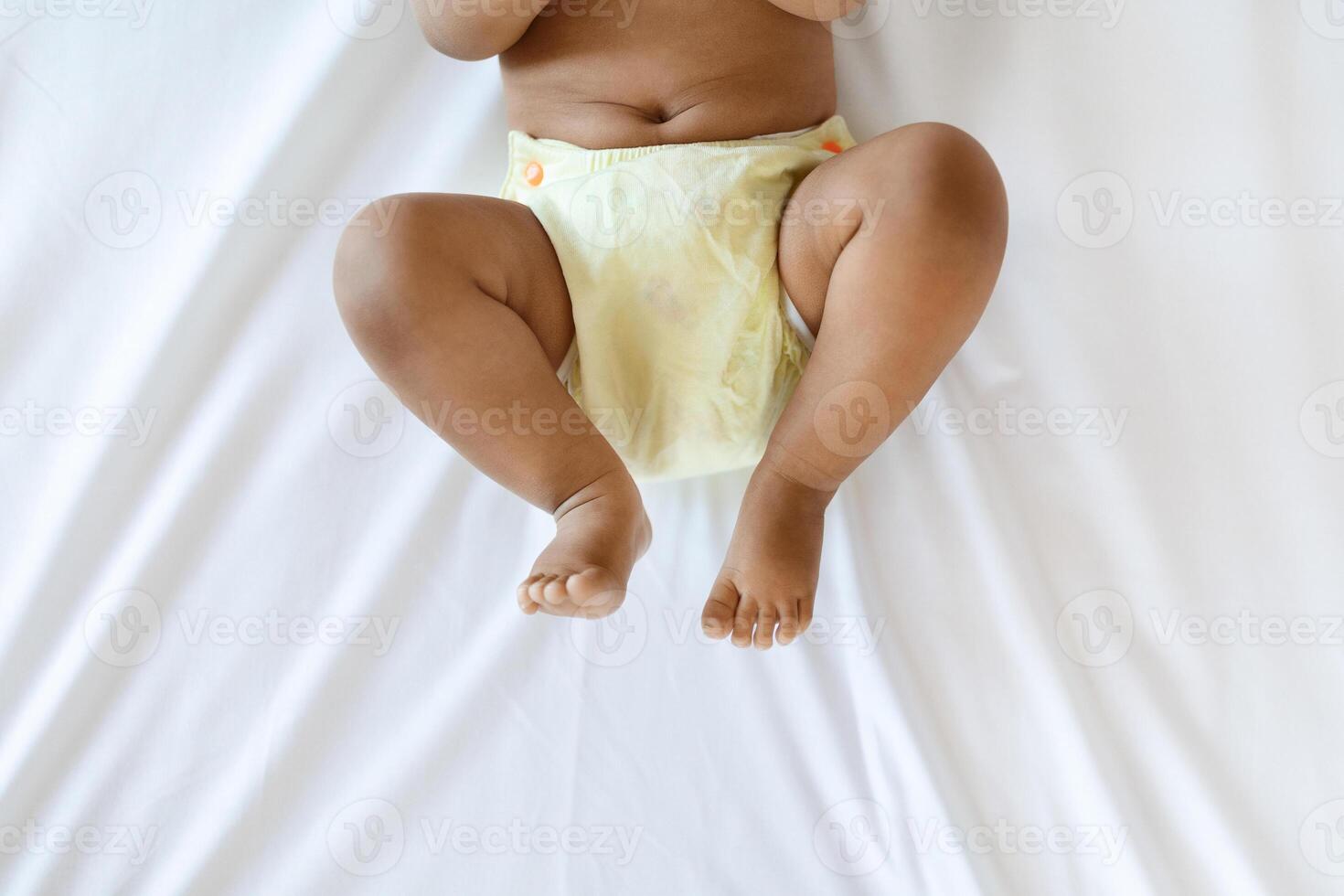 Cropped Shot Of Little African American Baby Wearing Reusable Diaper Lying On Bed, Bottom Part Of Unrecognizable Black Infant Boy Or Girl Resting On White Bedsheet In Bedroom, Top View photo