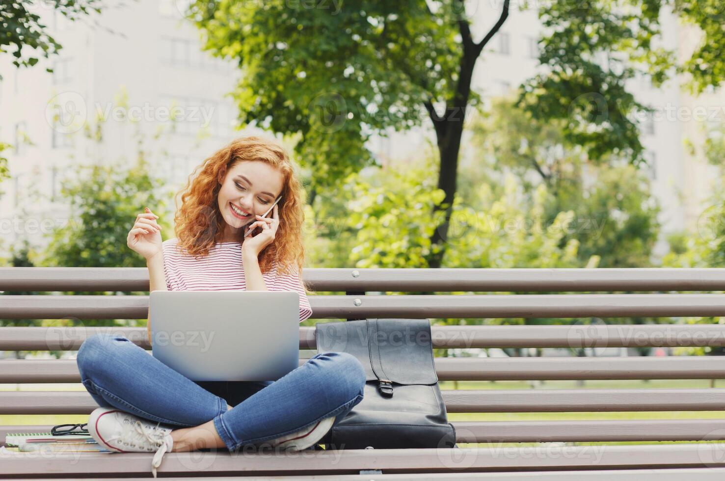 Young woman sitting on bench, talking on smartphone, working on laptop outdoors. Technology, communication, education and remote working concept, copy space photo