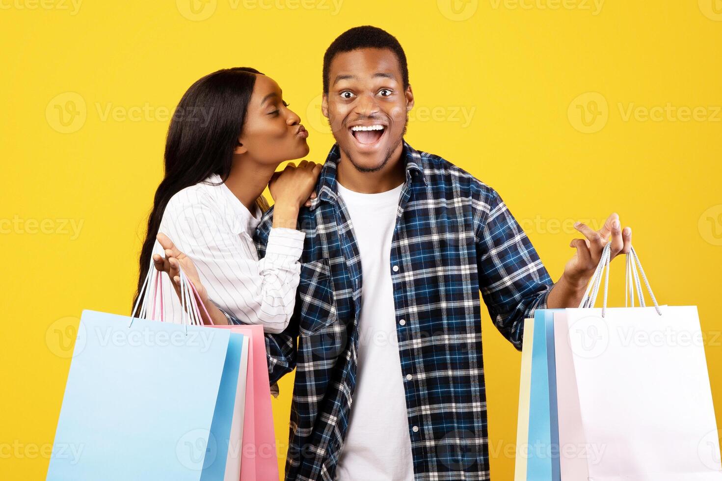 Seasonal sale and great offer. Surprised young african american guy with open mouth shows many color packages, lady kissing him on cheek, isolated on yellow background, studio shot, copy space photo