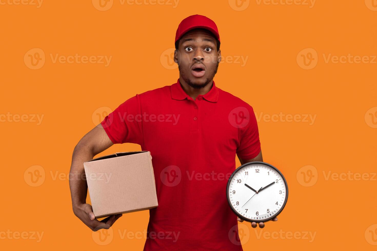 Express Delivery. Portrait Of Funny Surprised African American Male Courier In A Hurry Holding Cardboard Box And Clock Delivering In Time Standing Isolated Over Yellow Orange Background. Studio Shot photo