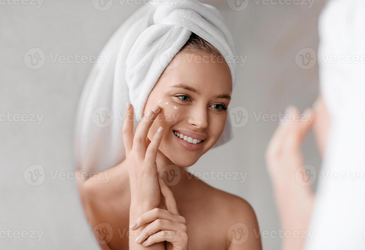 Portrait of young lady applying nourishing eye cream after shower, looking at mirror and smiling in bathroom interior. Anti-aging cosmetics, tools against lines, wrinkles eye care concept photo