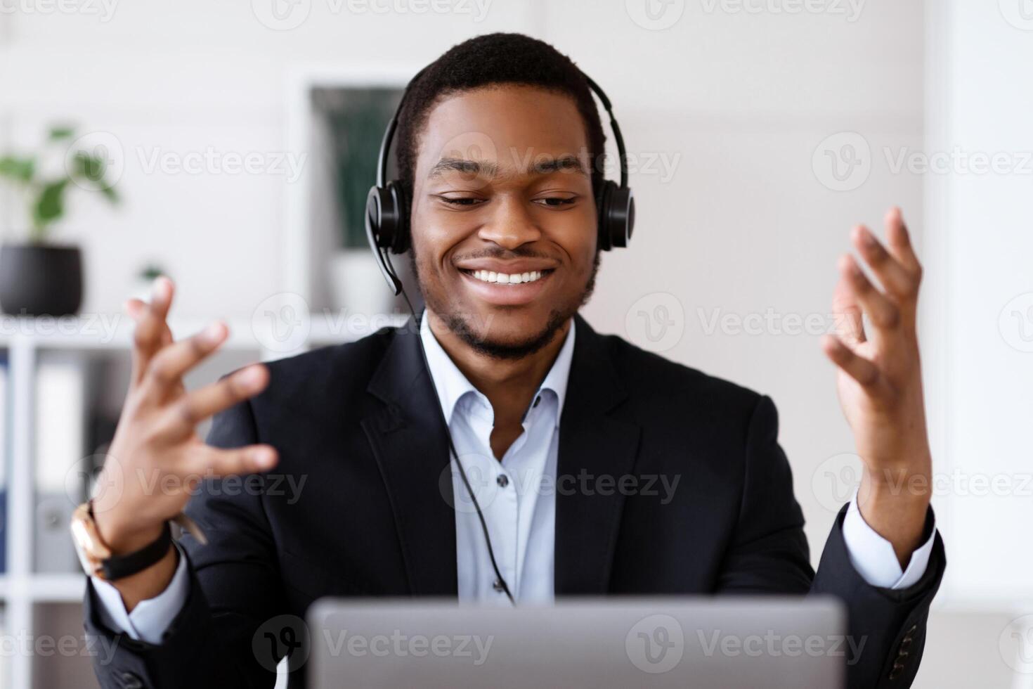 Closeup of inspired black businessman with headset having online conference, using laptop, talking to client or business partner, smiling at computer screen, business coach broadcasting from office photo