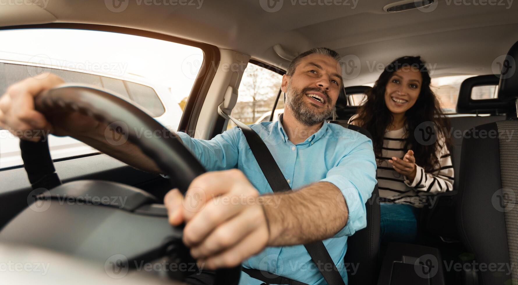 Portrait of happy male driver riding car, cheerful beautiful lady sitting inside auto on back passenger seat, looking on the road abd talking with guy, enjoying safe ride, windshield view photo