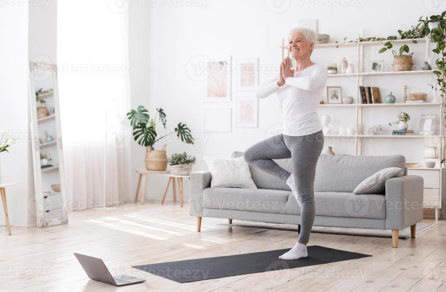 Sporty Senior Woman Practicing Yoga With Online Tutorials At Home, Standing In Tree Pose And Smiling, Enjoying Retirement Time, Free Space photo