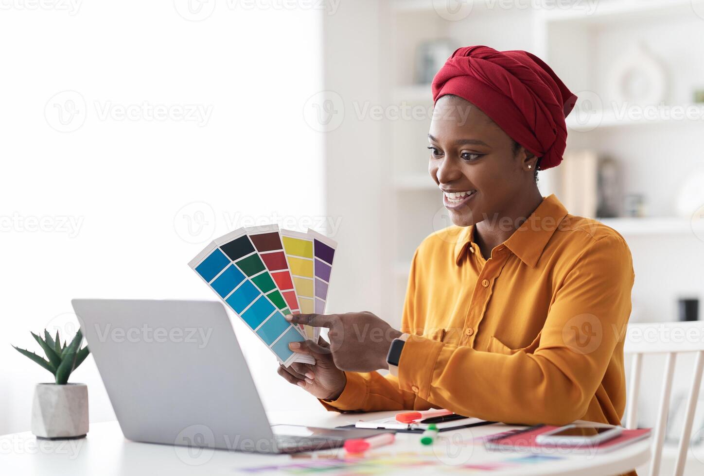 Stylish young black woman in red turban independent contractor having conference with client, female designer sitting at workdesk in front of laptop at home, showing color palette, copy space photo