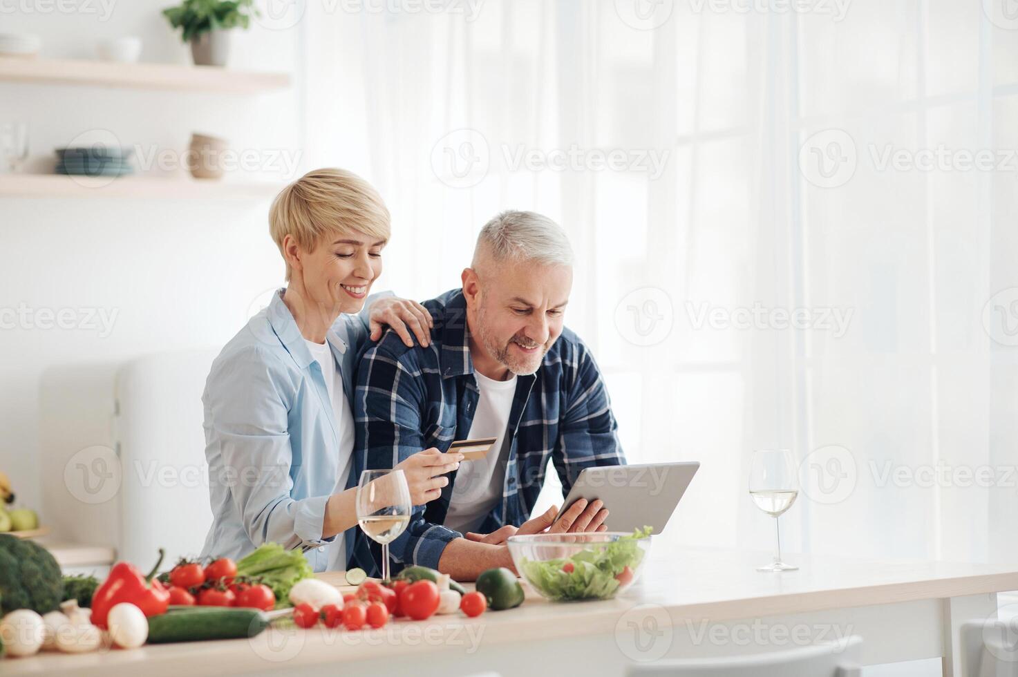 Healthy eating in middle age, paying bills, ordering groceries and shopping online. Smiling senior couple use laptop and credit card in modern comfort kitchen interior with bright colored vegetables photo