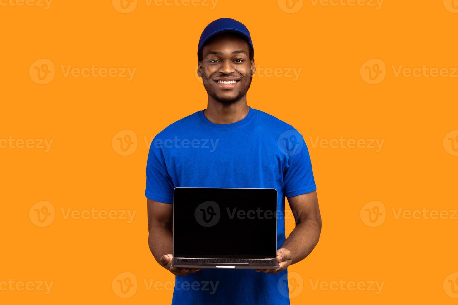 Track Your Order. Portrait of smiling black delivery man holding laptop with blank screen for mockup, wearing blue cap and uniform, showing computer with empty monitor isolated on orange background photo