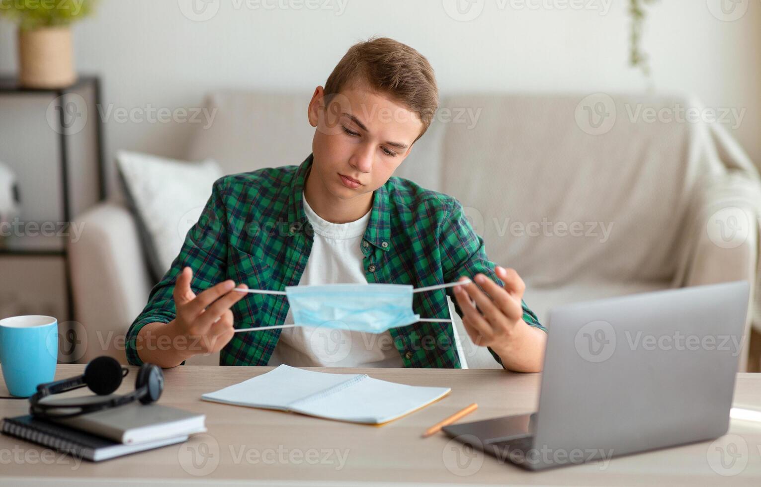 Pensive bored teen guy holding protective mask, studying at home during lockdown, empty space photo