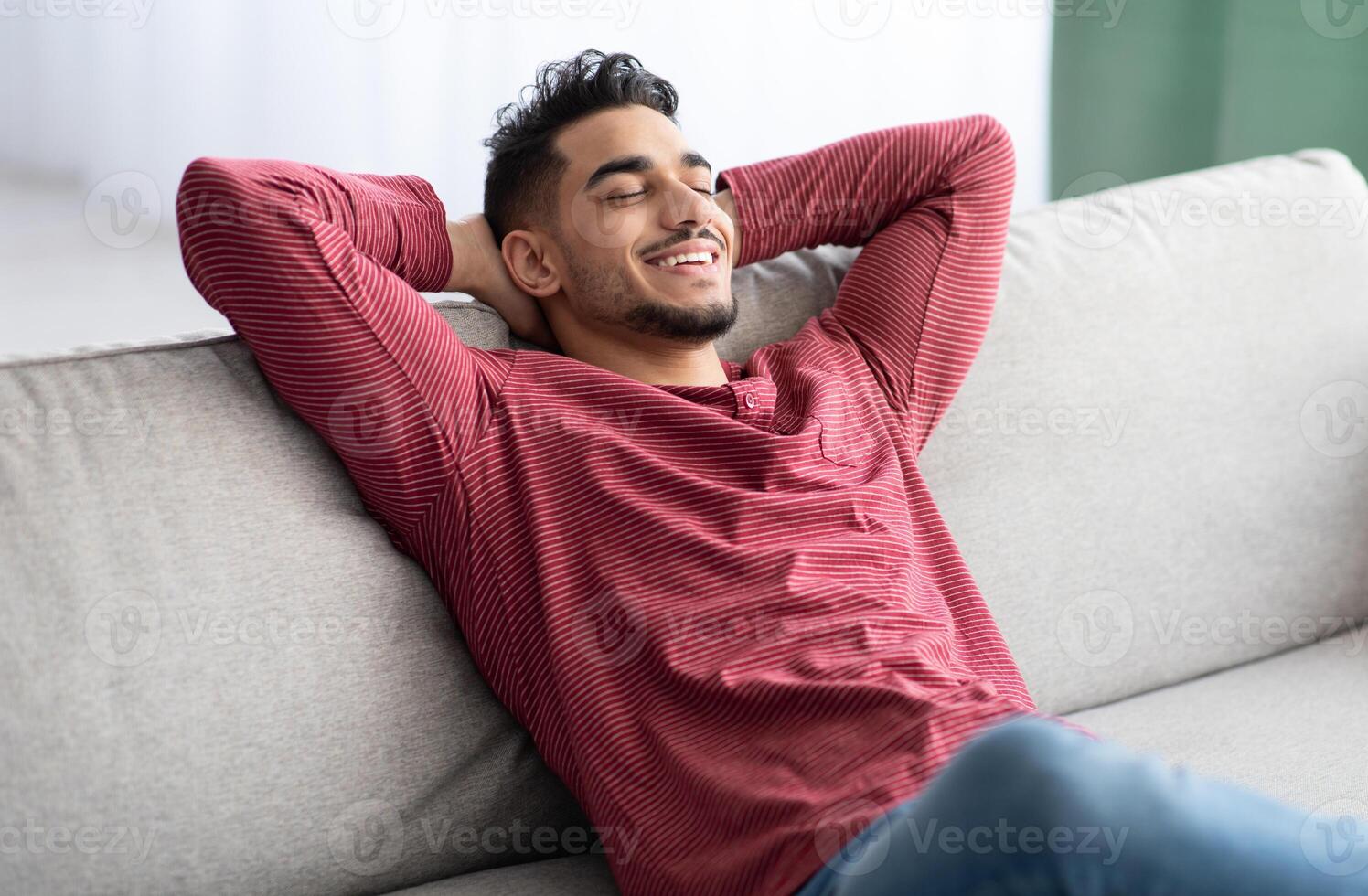 Happy middle-eastern guy in casual chilling at home, reclining on couch with closed eyes and smiling. Relaxed young arab man enjoying his new comfortable couch, leaning back, copy space photo