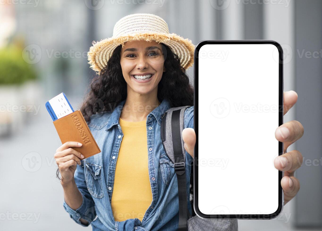 Happy attractive millennial brunette lady in straw hat tourist holding passport with flight tickets and cell phone with white blank screen, showing nice mobile app for travellers, mockup photo