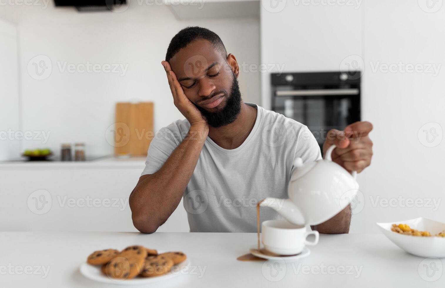 Lack Of Sleep Concept. Portrait of tired young African American man sleeping while sitting at dining table in kitchen, holding kettle pouring coffee away from cup spilling hot drink on desk photo