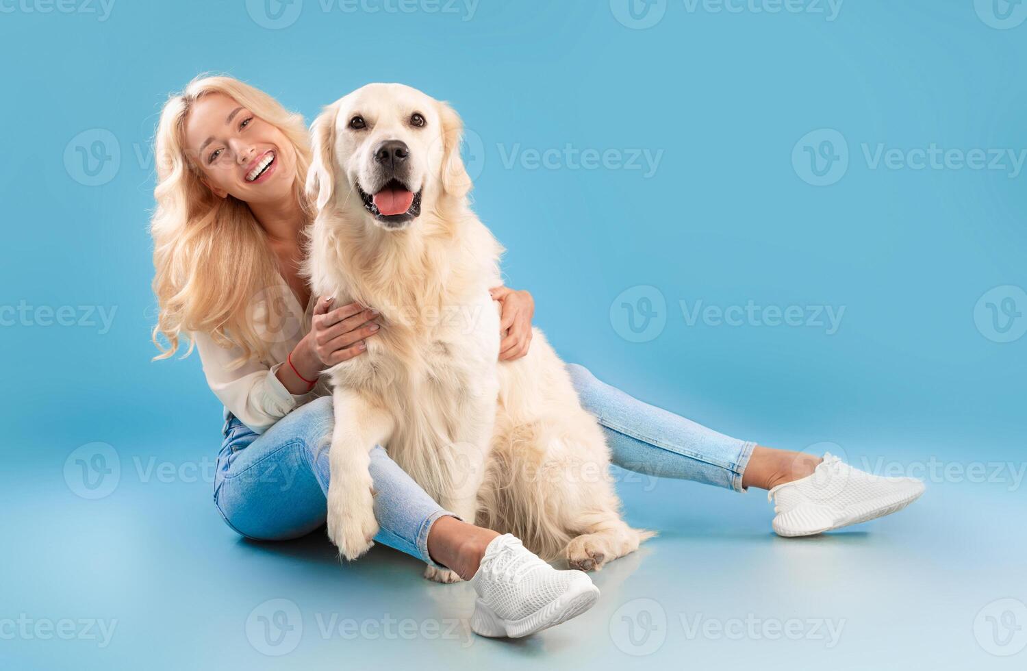 Love And Care Concept. Cheerful young lady patting her happy labrador spending time together, embracing pet, posing looking at camera sitting on the floor isolated on blue background, full body length photo