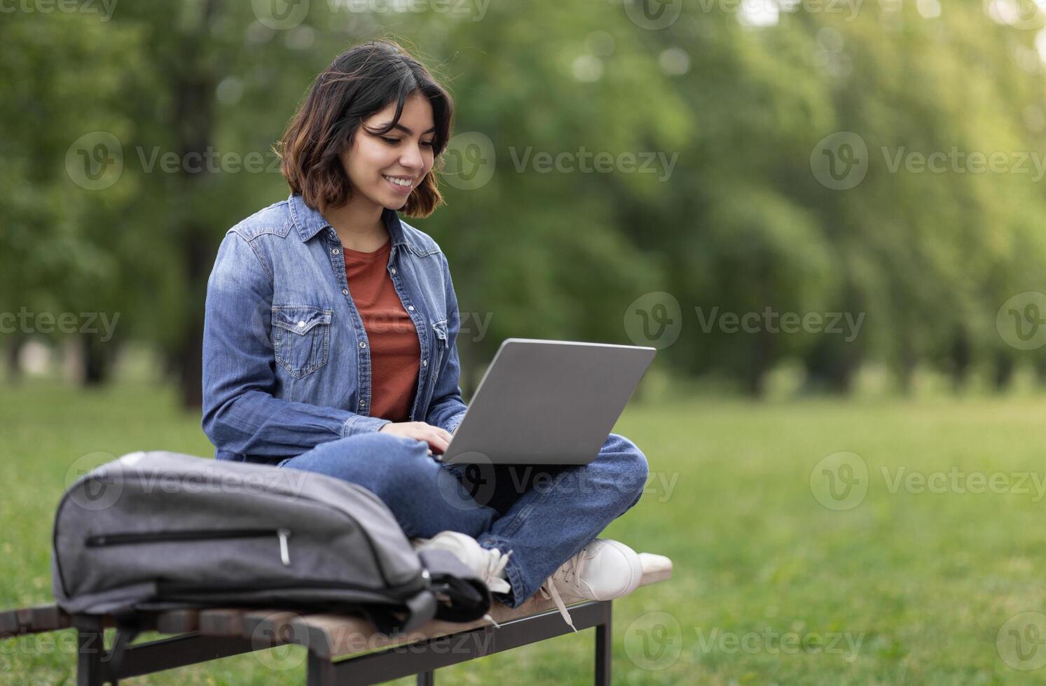 Distant Learning. Young Arab Female Student Using Laptop While Sitting On Bench Outdoors In Park, Smiling Middle Eastern Woman Watching Educational Webinar, Enjoying Remote Study, Copy Space photo