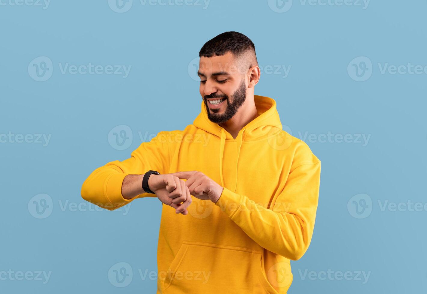Young arab guy checking his smartwatch on blue studio background. Happy middle eastern man using fitness tracker. Modern technologies and active lifestyle concept photo